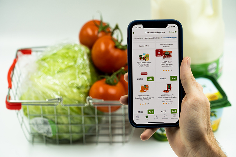 A mobile phone showing online grocery shopping apps with a basket of different groceries in the background.