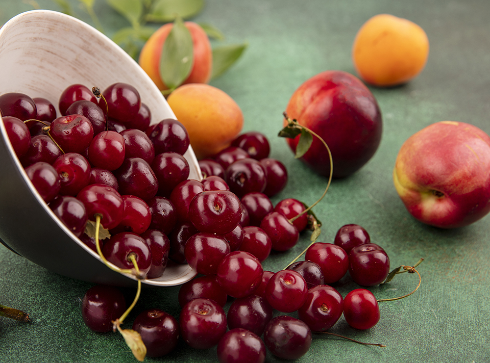 Side view of cherries spilling out of bowl with apricots and peaches on green background. Copyright: Stockking/Freepik.